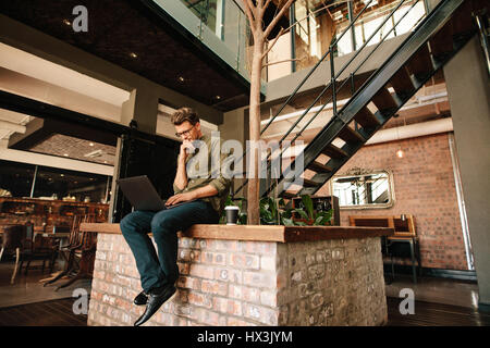 Thoughtful young man sitting in office cafeteria and looking at laptop. Executive using laptop during coffee break. Stock Photo