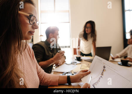 Close up of young woman looking at document during meeting in conference room. Businesswoman reading business chart with people in background. Stock Photo