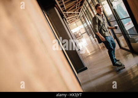 Full length of a handsome young man skateboarding in office corridor. Casual businessman skating in modern office. Stock Photo