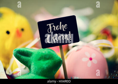 a chalkboard with the text frohe ostern, happy easter in german written in it and a pile of decorated easter eggs of different colors, a green rabbit  Stock Photo