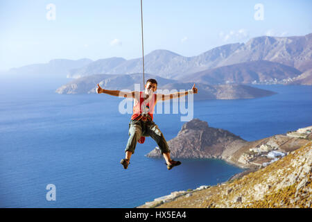 Cheerful rock climber swinging on rope and showing thumbs up against view of coast Stock Photo