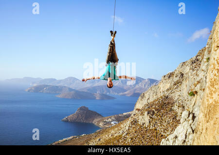 Cheerful rock climber swinging on rope upside down while fooling around, against view of coast Stock Photo