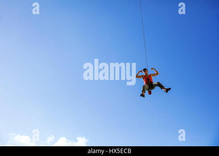 Rock climber swinging on rope and flexing muscles jokingly against blue sky Stock Photo