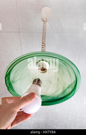 Replacing the light bulb in the ceiling light LED lamp in human hand, next to the lampshade of green frosted glass. Stock Photo