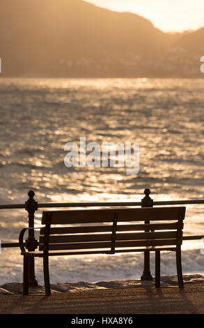 Wooden bench on the promenade of the popular resort town of Llandudno in North Wales as the sun begins to rise over the little orme along the coast Stock Photo