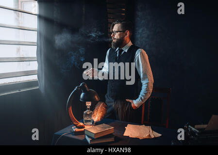 Thoughtful bearded writer in glasses smoking a cigarette and look at the window with sunlight. Retro typewriter, feather, crystal decanter, books and  Stock Photo