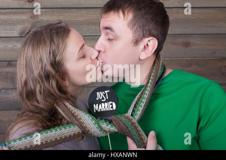 Love couple kisses wrapped in a scarf and shows funny icon on a stick with just married inscription, wooden background. Fun photo props and accessorie Stock Photo