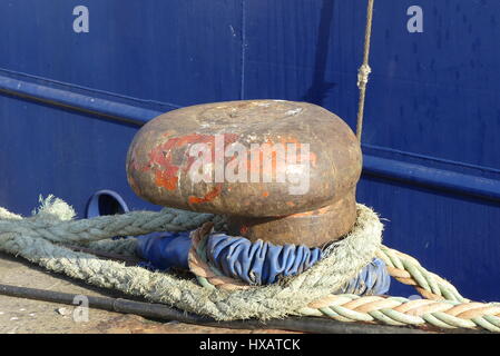 Closeup of rusty bollard with mooring ropes on the  dock with blue ship hull on background Stock Photo