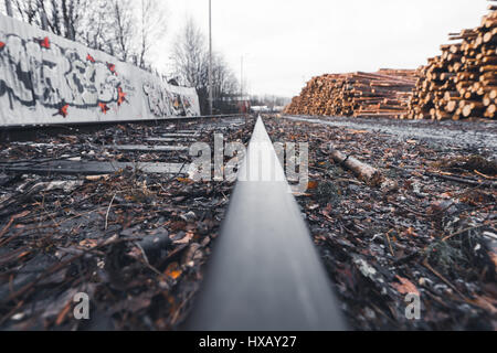 Wide and low angle view of railroad track covered by debris dividing blurred metal wall and pile of wood on overcast and wet day Stock Photo