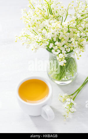 Cup of tea and bouquet of Lilies of the Valley in glass vase on rustic table. Stock Photo