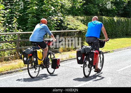 Couple cycling English road on cycle route for bicycle along quiet country lane in Essex England UK in summer sunshine with bike panniers fitted Stock Photo