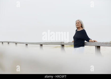 Confident senior woman leaning on beach boardwalk railing Stock Photo