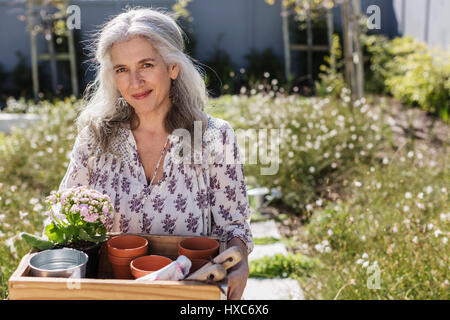 Portrait mature woman carrying gardening tray in sunny garden Stock Photo