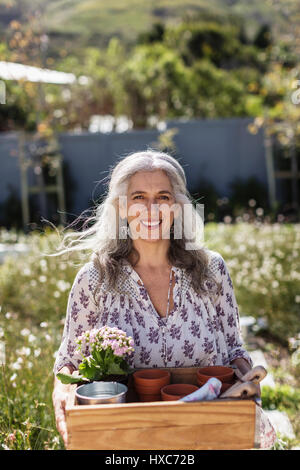 Portrait smiling mature woman carrying gardening tray in sunny garden Stock Photo