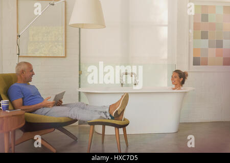 Husband with digital tablet relaxing, talking to wife in soaking tub in luxury hotel room Stock Photo