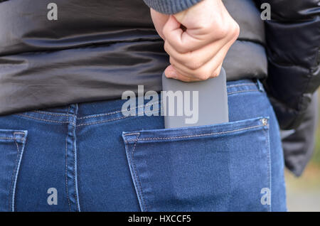 Pickpocket stealing a mobile from a vulnerable woman stealthily pulling it out of the rear pocket of her jeans outdoors in a public place Stock Photo