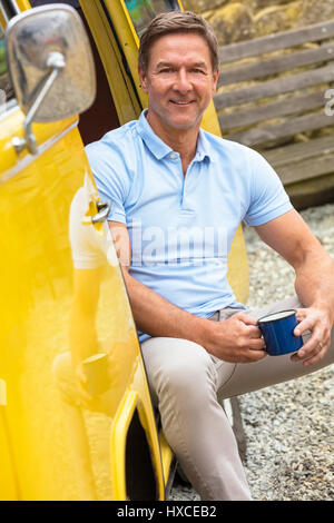 Handsome, successful and happy middle aged man sitting in the doorway of a yellow camper van bus drinking tea or coffe form a tin cup or mug