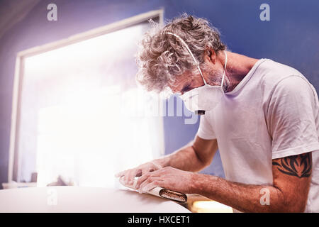 Focused man wearing protective mask sanding surfboard in workshop Stock Photo