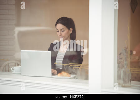 Young businesswoman using laptop in coffee shop seen through window Stock Photo