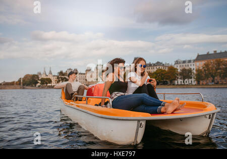 Shot of teenage girls sitting in front of a pedal boat and using mobile phone with men pedaling the boat in background. Young people enjoying boat rid Stock Photo