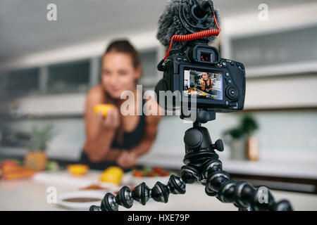 Young woman recording video on tripod mounted camera in kitchen. Camera showing woman with an orange in hand. Stock Photo