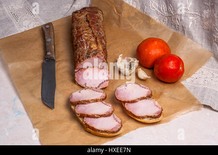 Slices smoked with spice and herbs meat or ham and old vintage knife on brown packing paper. Tomatoes, garlic and two small snacks of bread and meat a Stock Photo