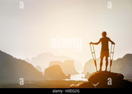 Disabled man with crutches stands on a big rock and looking at great view to islands and mountains. Space for text. Winner pose Stock Photo