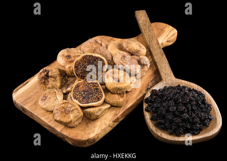 Dried figs on wooden chopping board and black raisins in wooden spoon on black background Stock Photo