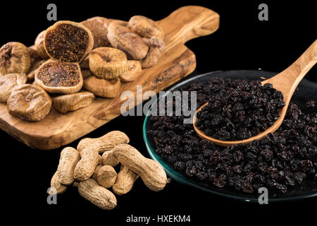Dried figs on wooden chopping board, peanuts and black raisins in plate on black background Stock Photo