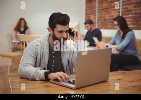 Male executive talking on mobile phone while using laptop at desk in office Stock Photo