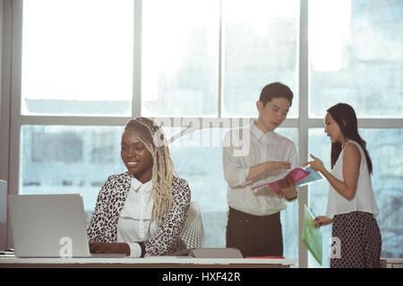 Female executive using laptop at desk against colleagues discussing in background Stock Photo