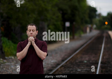 Man praying alone by train tracks with hands clasped together. Stock Photo