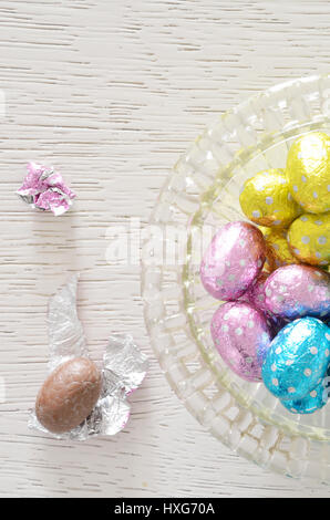Chocolate egg foil wrapper and chocolate egg beside a glass bowl with foil wrapped Chocolate on white wooden table Stock Photo