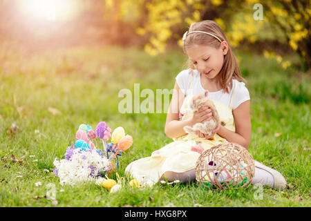 Beautiful smiling little girl holding cute bunny and sitting on the grass with Easter eggs in spring holidays. Stock Photo