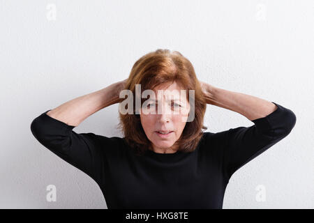 Upset senior woman in black t-shirt, holding head, having headache. Studio shot against white wall. Stock Photo