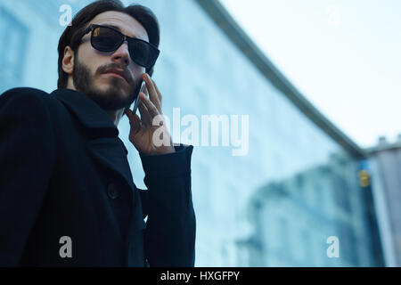 Low angle portrait of handsome modern man wearing sunglasses and black coat making phone call outdoors in streets of business city Stock Photo