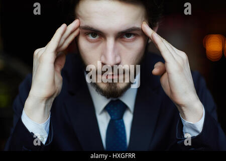 Head and shoulders portrait of handsome bearded man wearing elegant business suit, looking strongly at camera while rubbing his temples in thinking pr Stock Photo