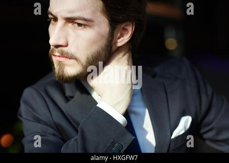 Sunlit head and shoulders portrait of handsome bearded man wearing elegant business suit, looking away frowning and rubbing his neck Stock Photo