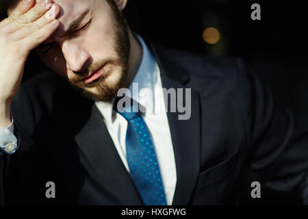 Sunlit head and shoulders portrait of handsome bearded man wearing elegant business suit, closing his eyes and rubbing forehead trying to relieve tens Stock Photo