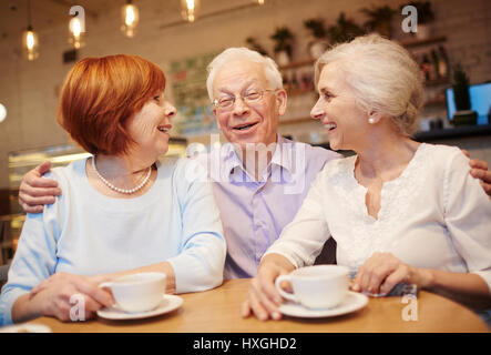 Senior man embracing two females while sitting in cafeteria and talking Stock Photo