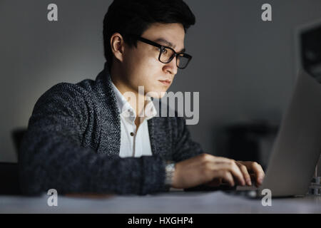 Side view portrait of focused Asian man wearing glasses working with laptop in dark room late at night, his face lit up by screen Stock Photo