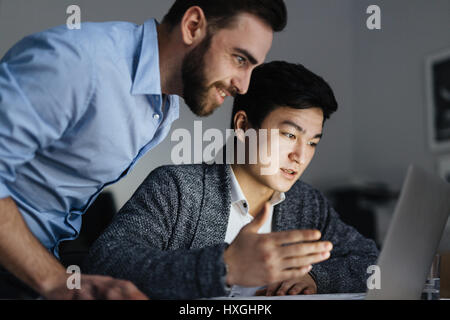 Portrait of two office workers, one of them Asian, collaborating on project and using laptop in dark room late at night, faces lit by lamp light Stock Photo