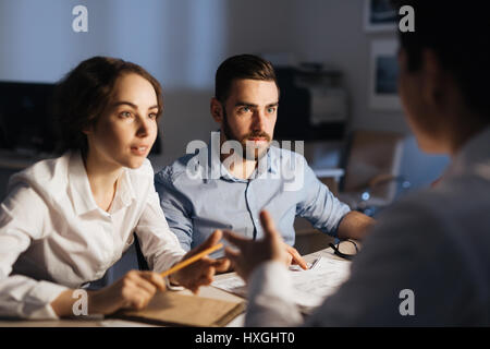 Portrait of three colleagues working overtime in dark office late at night: brainstorming at meeting table while collaborating on project trying to fi Stock Photo
