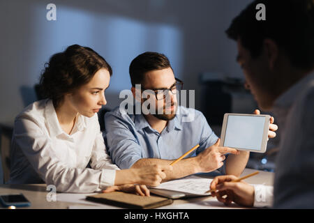 Portrait of three colleagues working overtime in dark office late at night: pointing at blank computer screen and explaining something while having li Stock Photo
