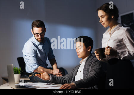 Portrait of three colleagues working overtime in dark office late at night: pointing at computer screen and discussing project while drinking coffee Stock Photo