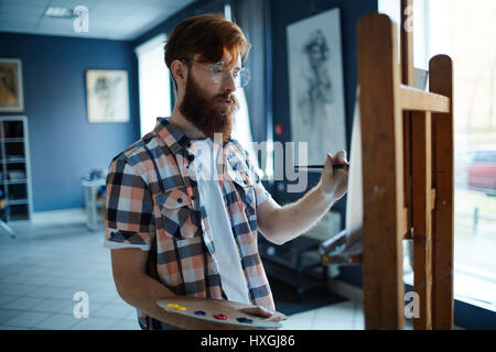 Young man with paintbrush and palette standing in front of easel in art-studio Stock Photo