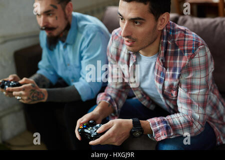 Portrait of two modern adult men, one of them heavily tattooed,  holding controllers and playing video game on couch, looking tense in competition Stock Photo
