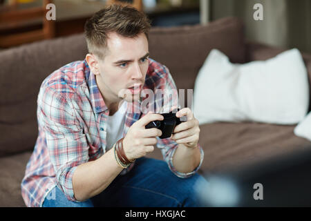 Portrait of emotional adult man playing video game holding wireless controller while sitting on couch at home Stock Photo