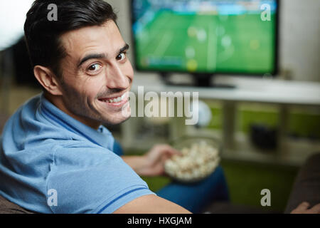Portrait of handsome man smiling, turning to look at camera while watching football match at home with popcorn Stock Photo