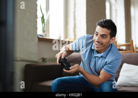 Portrait of handsome emotional adult man playing video game holding wireless controller and  smiling joyfully while sitting on couch at home Stock Photo